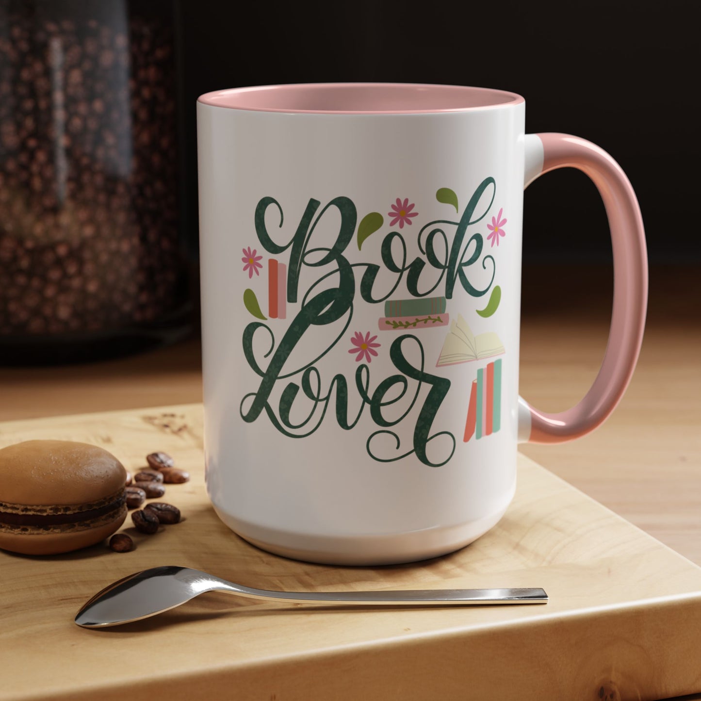 White mug with 'Book Lover' design on a wooden surface with coffee beans and a macaron.