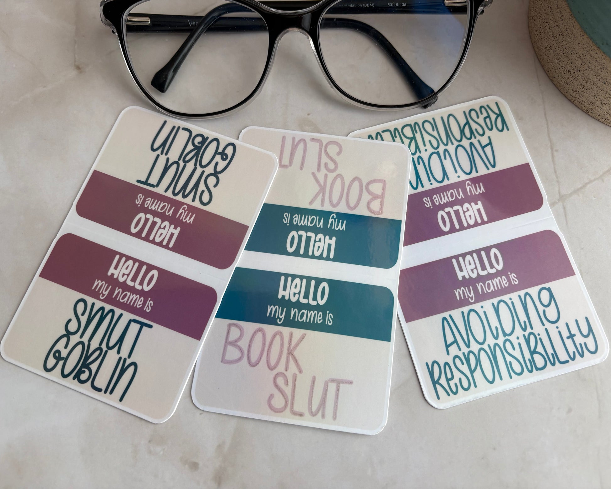 Three name tag magnetic bookmarks with text on a marble surface with glasses in the foreground.